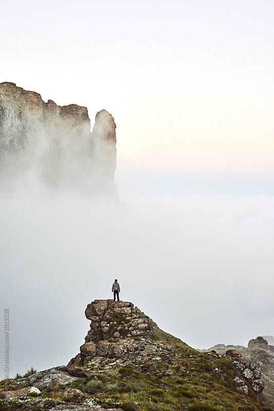 From a precarious and airy rock peak a hiker stands in owe of the majestic misty mountains that surround him.