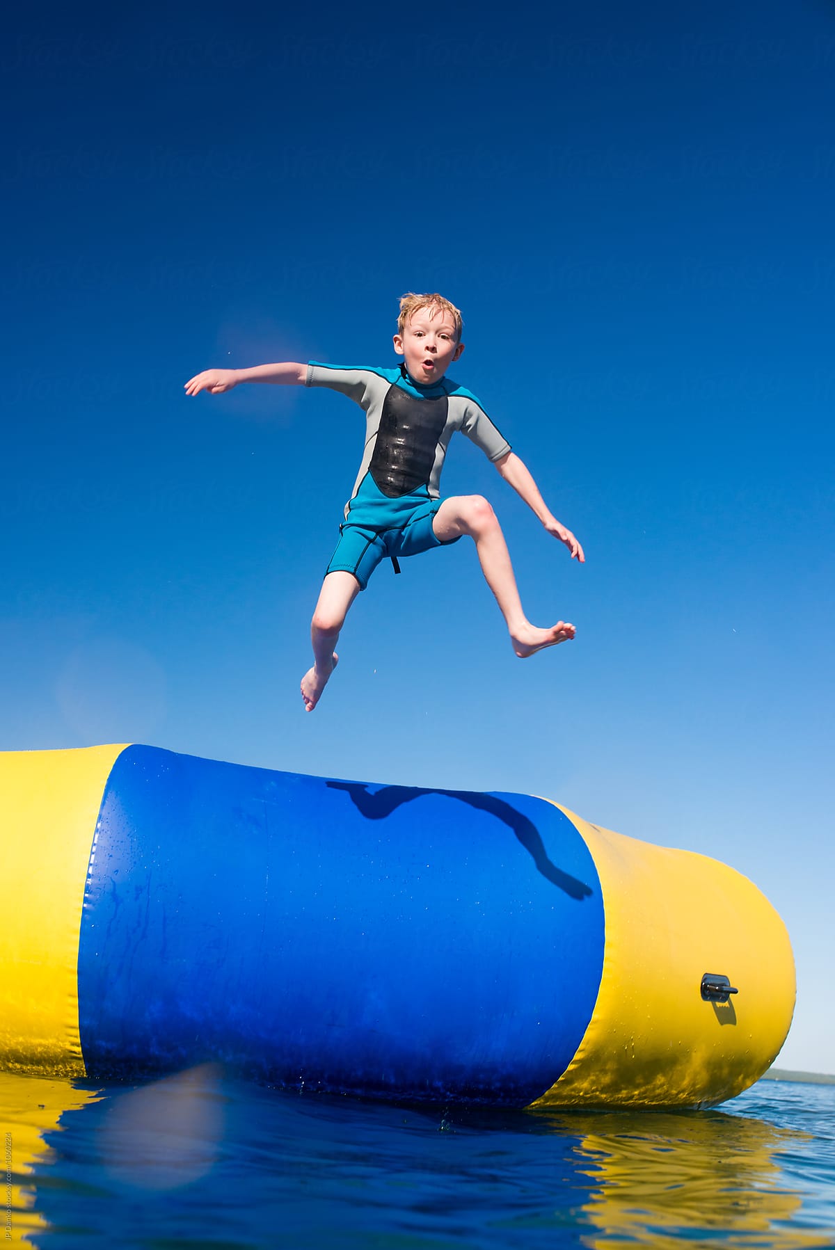 Boy Jumping Into Summer Lake From Water Trampoline At Cottage on Sunny Summer Day