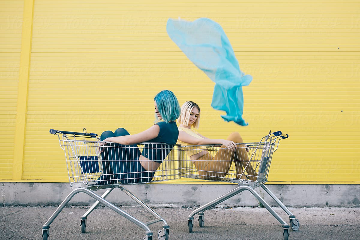 Two Female Friends Sitting In Front Of A Yellow Wall In A Grocery Cart By Stocksy Contributor