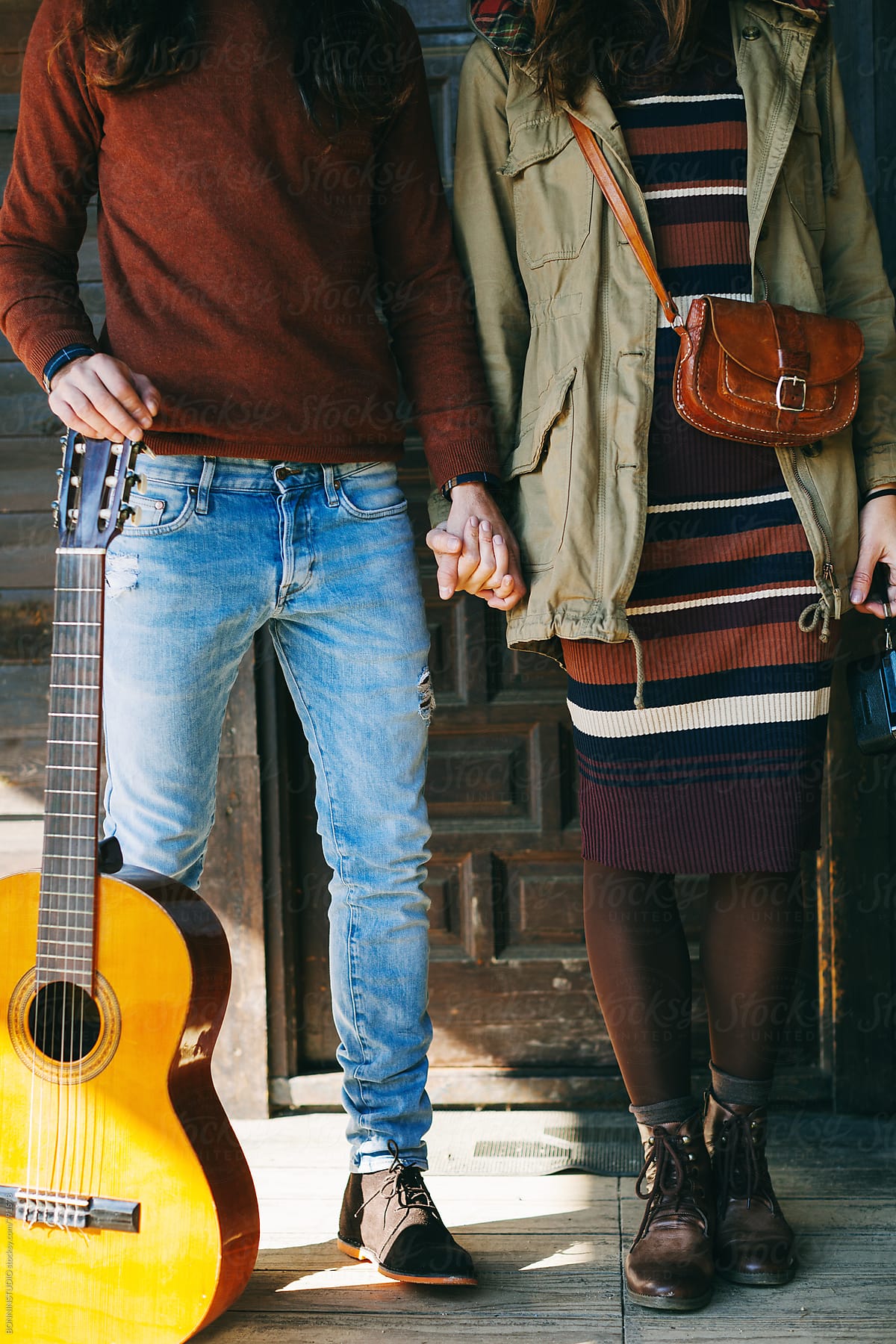 Closeup of hipster couple with a guitar holding hands on a cabin porch.