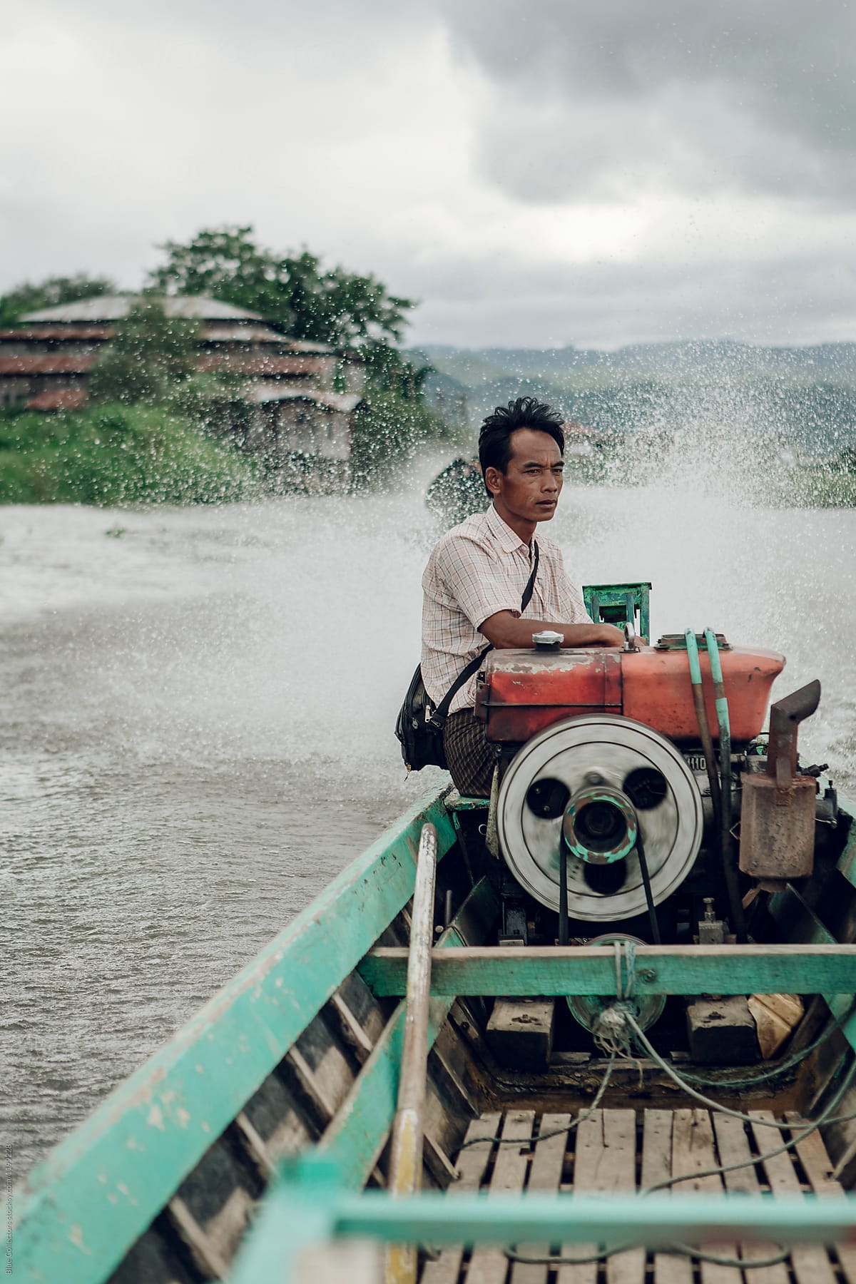 Asiatic handsome man seated and driving small motor boat with village in the background