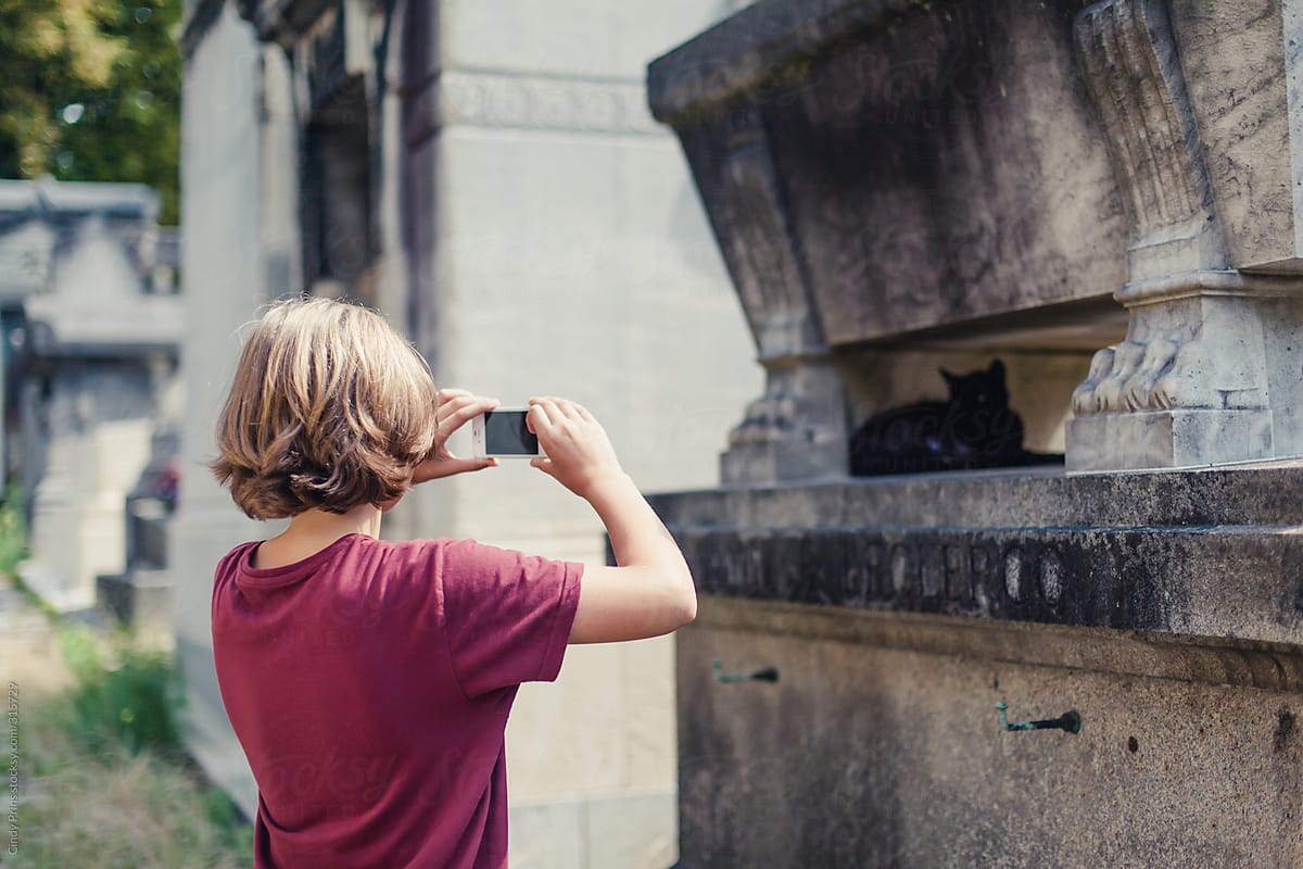 Boy photographing a black cat on a grave in a cemetery in Paris with his smartphone