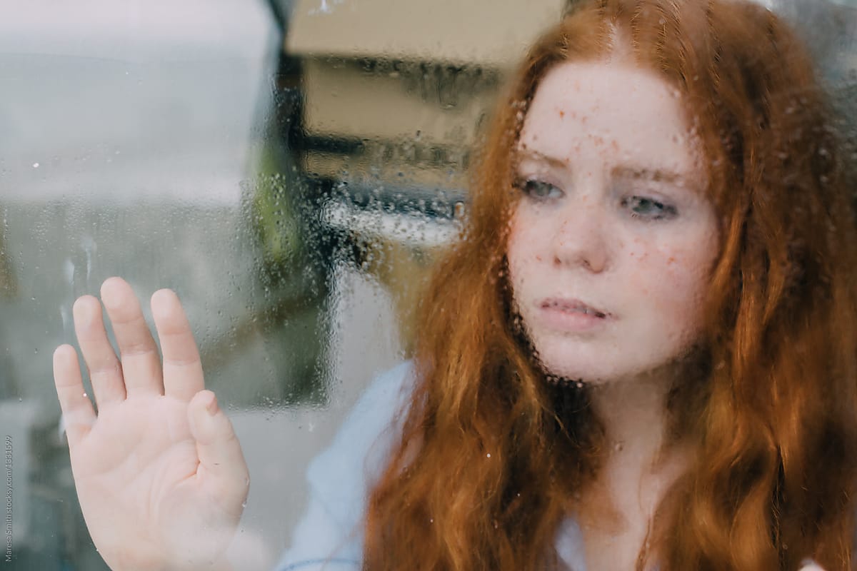 A ginger-haired teenager holding her hand against a rainy window, looking pensive