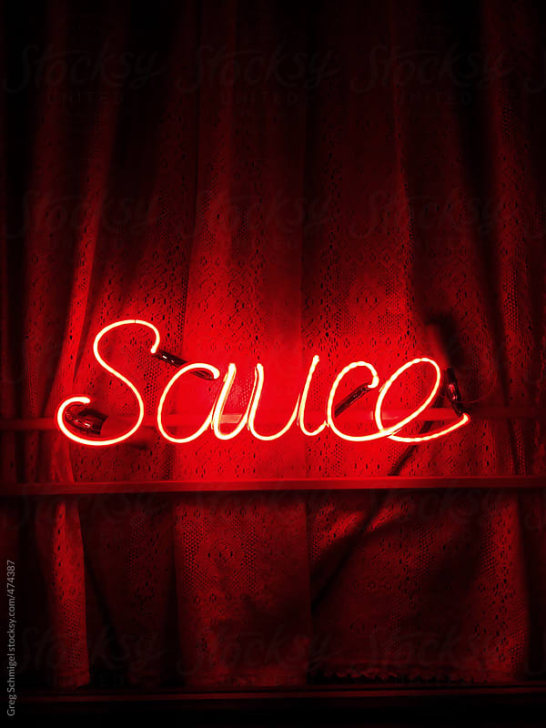 The word sauce illuminated in a red neon sign, hanging in front of a sexy red curtain