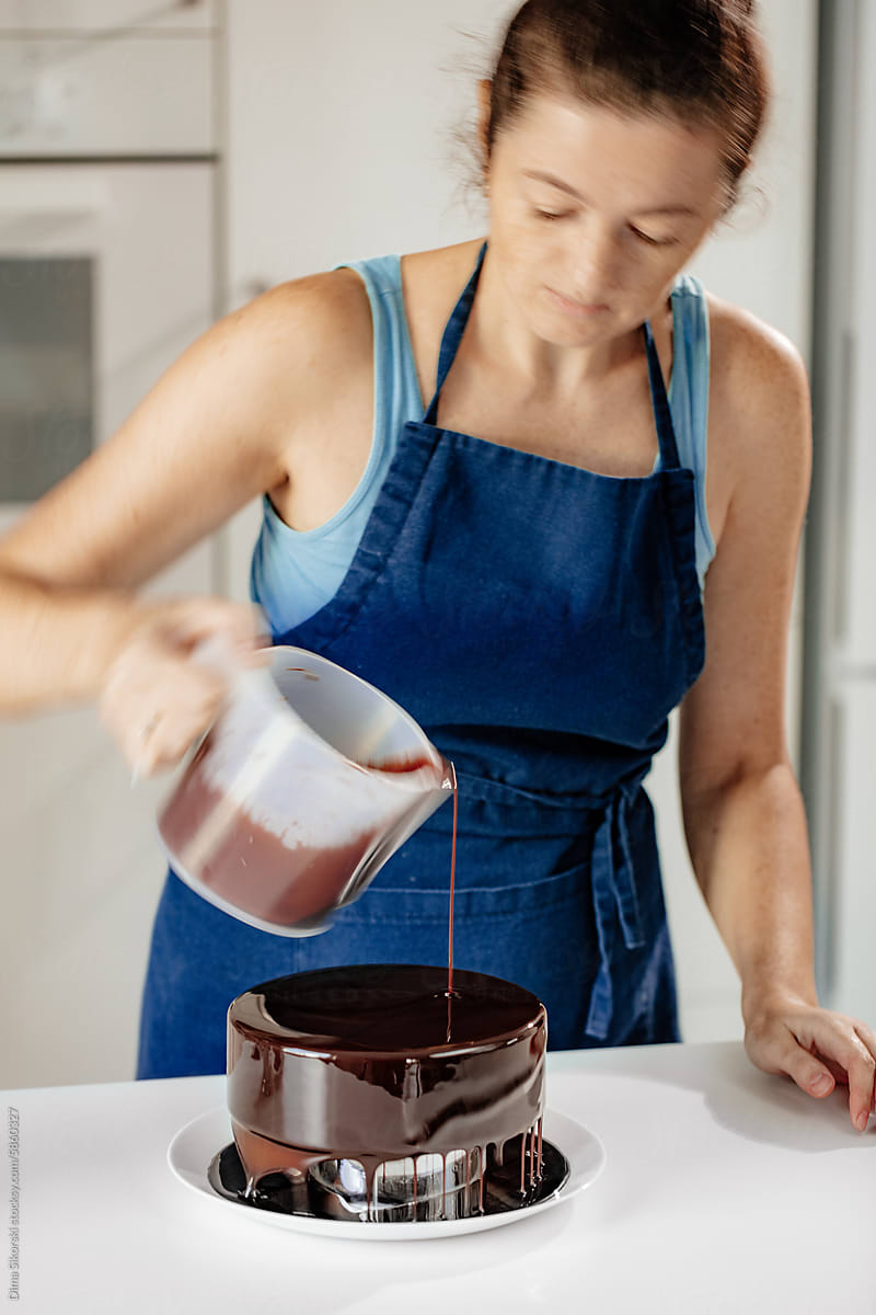 A female in an apron pours chocolate icing on the cake
