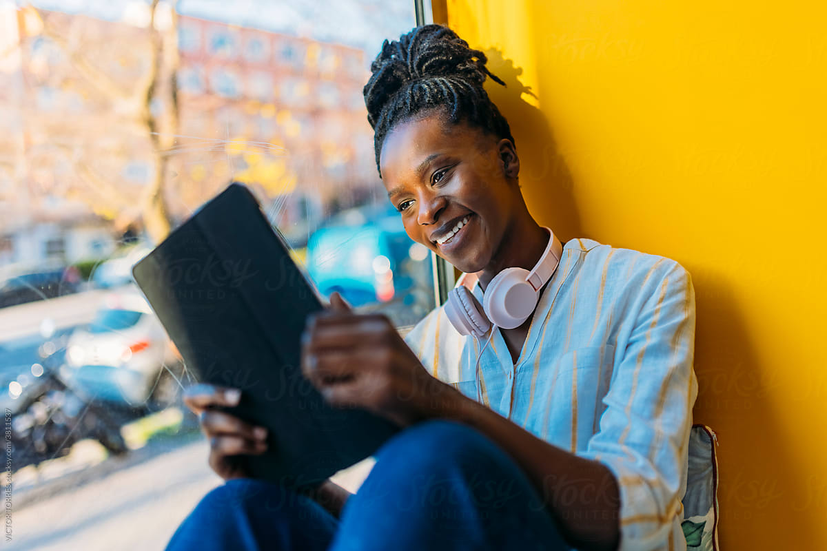 Black woman browsing tablet sitting on windowsill
