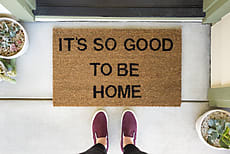 Woman standing in front of a doormat reading âItâs so good to be homeâ by Amy Covington for Stocksy United