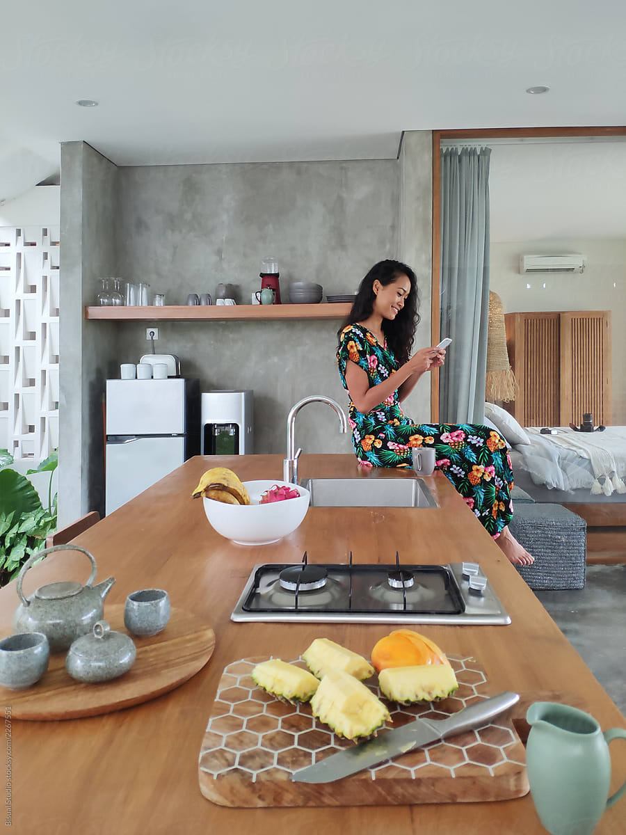 Asiatic smiling woman texting with cell phone in kitchen