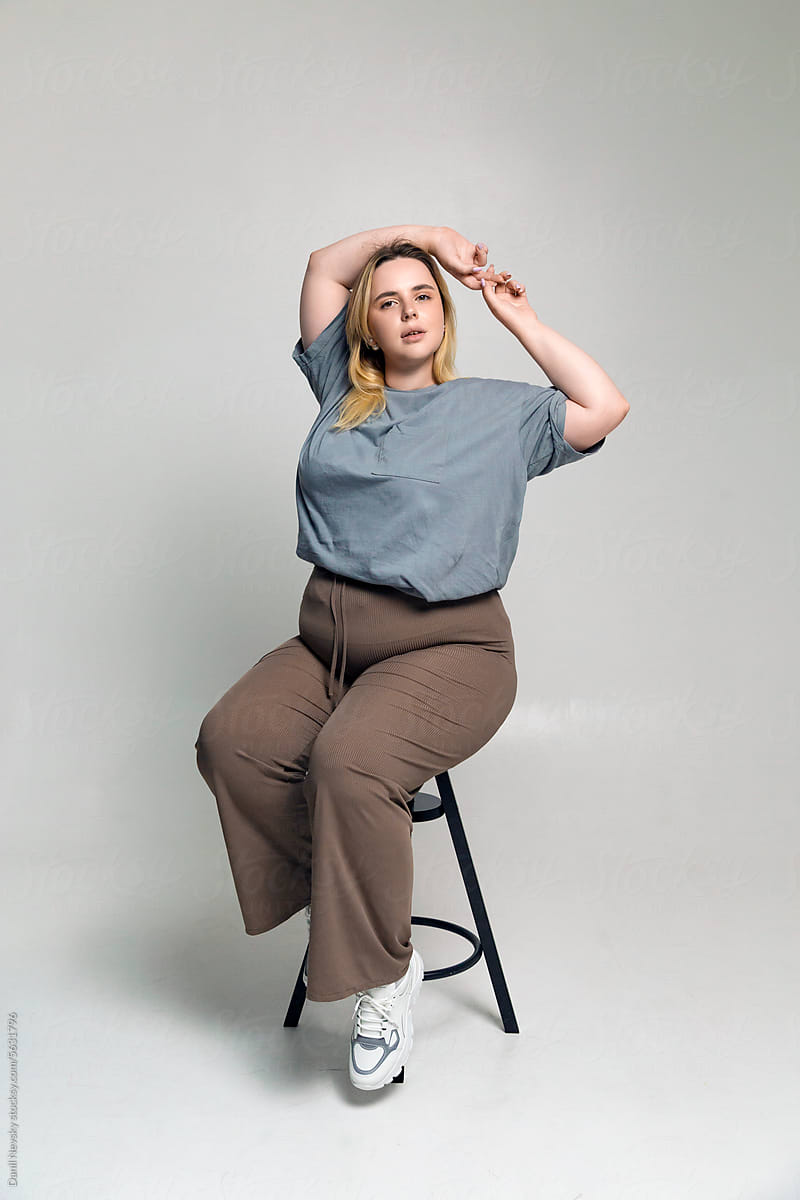 Young plus size woman sitting on stool in room with gray backdrop