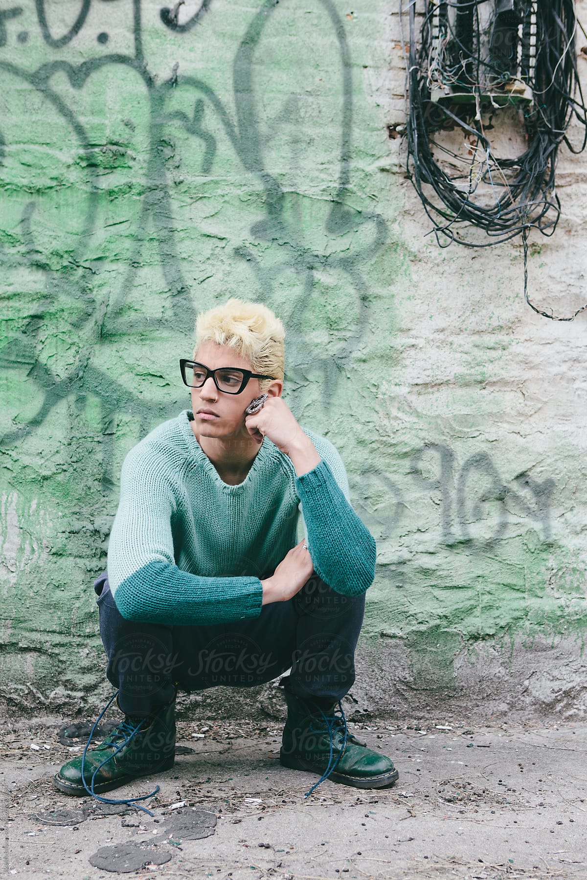 Stylish Young Man with Bleached Blond Hair in Spring and Summer Fashion Squatting against Graffiti Wall