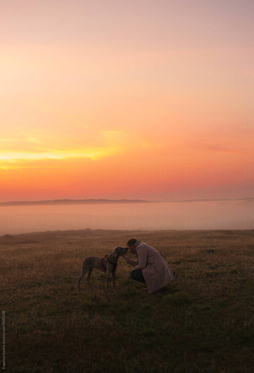 A young man squatting in front of the dog and  holding the dog\'s face in the middle of the field in the foggy morning with an orange red sky