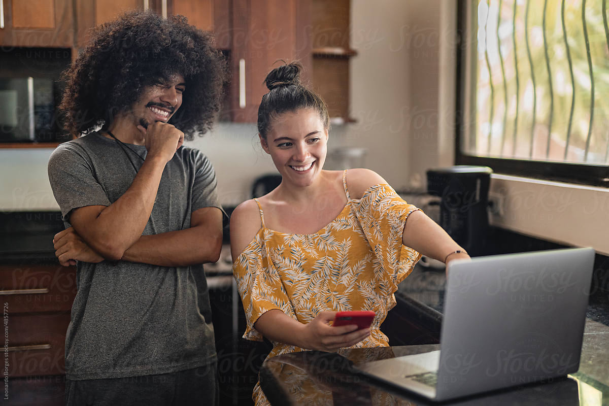 Couple in their home kitchen using a cell phone and laptop computer