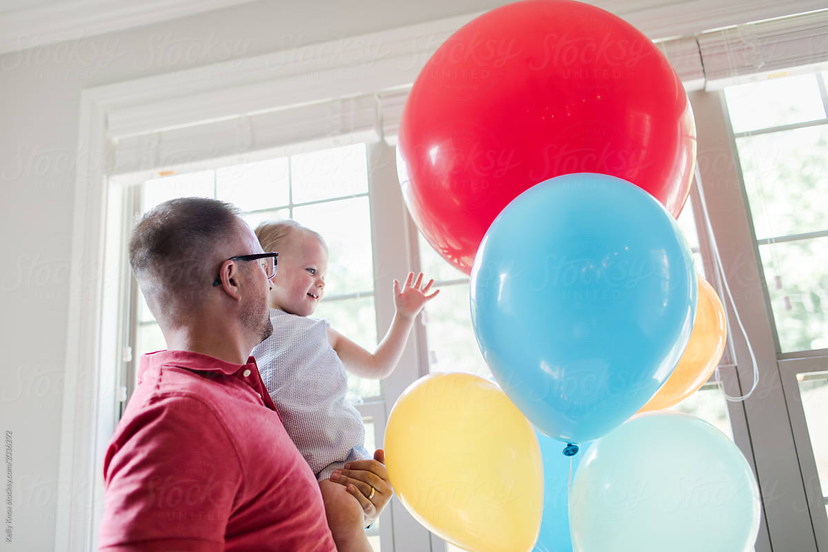 father holding his toddler up to touch birthday balloons