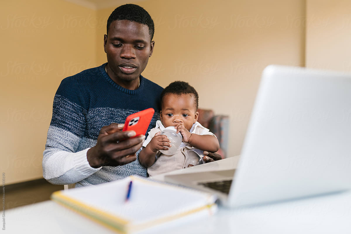 Black father with baby using mobile phone during work