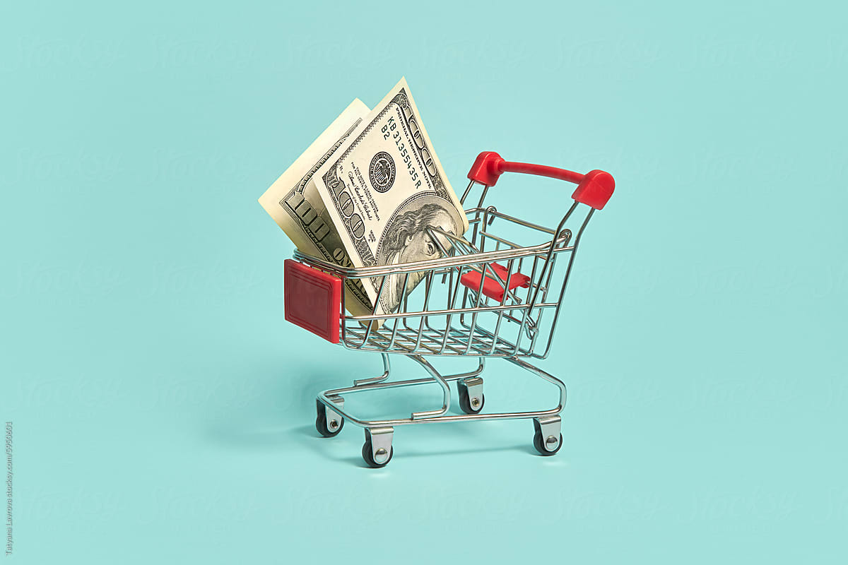 Mini shopping cart with dollar bills on light blue background.