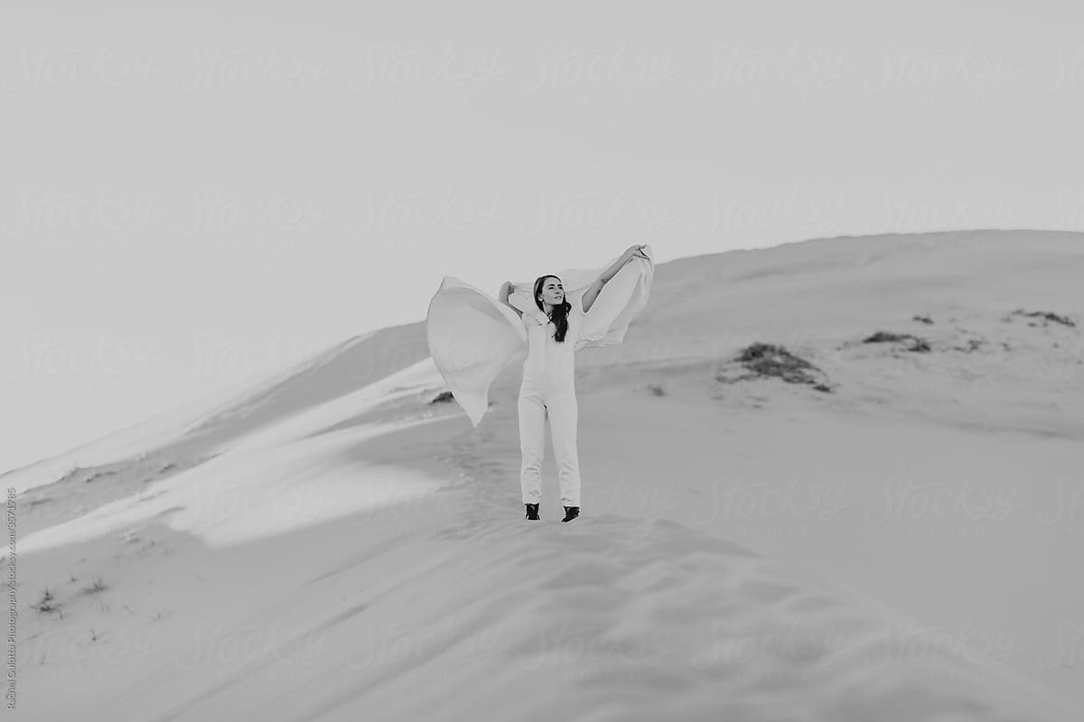 Girl in White Jumpsuit and Black Boots with White Sheet Standing on a White Desert Dune