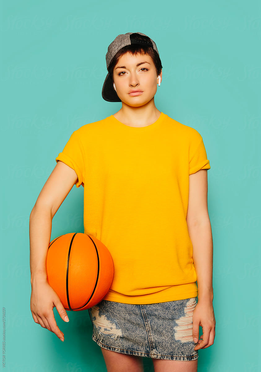 Portrait of cute girl in sporty outfit over blue background holding a basketball ball.