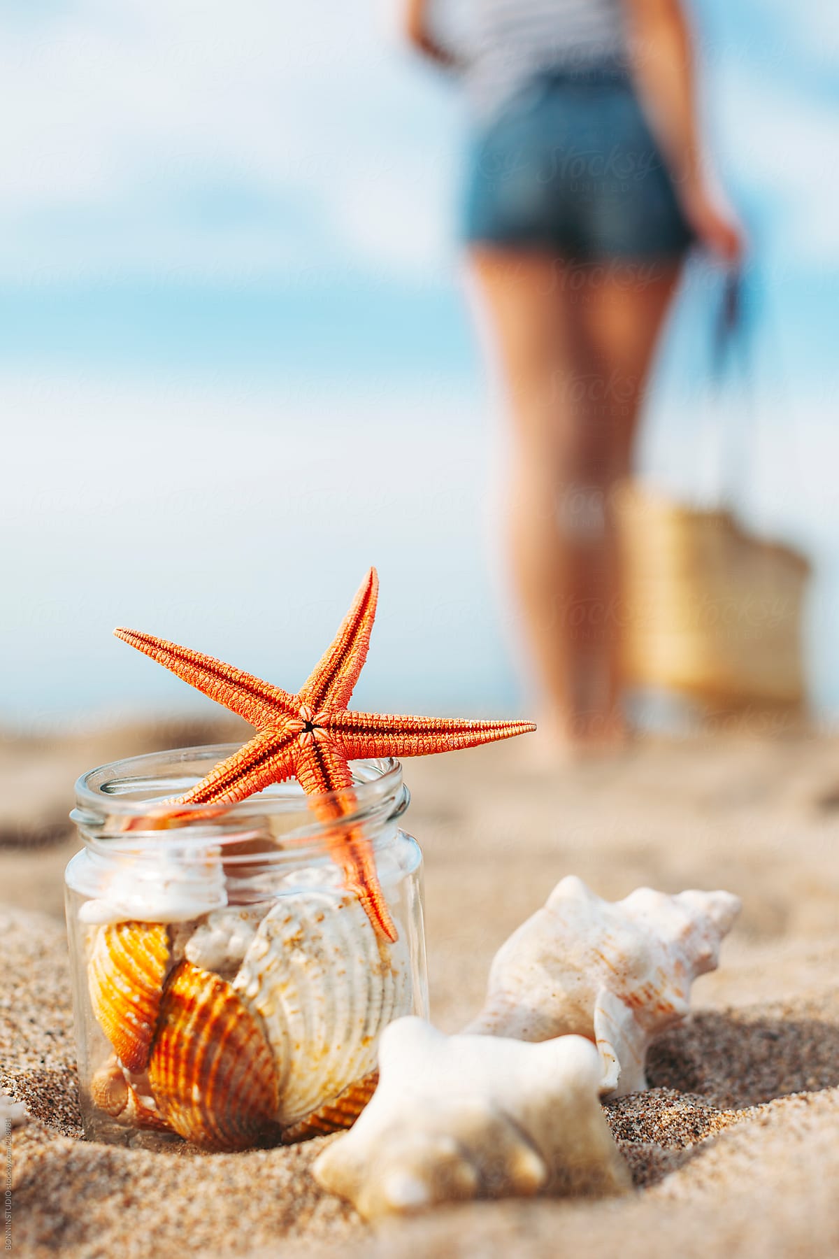 Starfish and shells inside a pot on the beach. Woman standing with straw basket on the background.