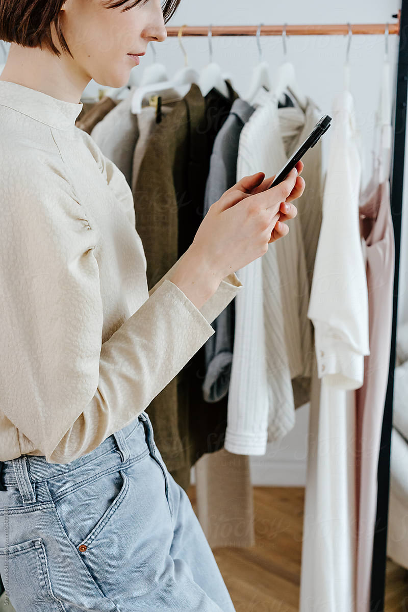 Crop female browsing smartphone near rack with clothes