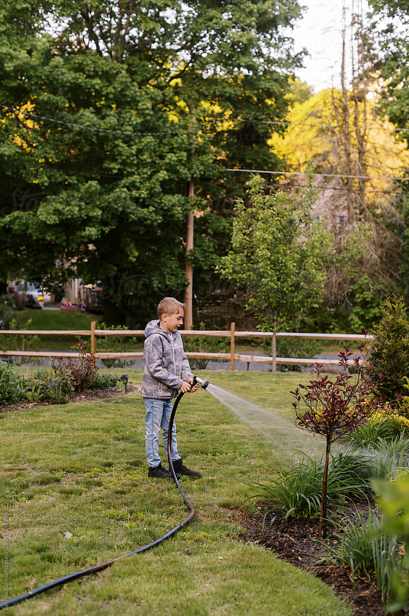 boy standing in garden and watering plants in the evening with hose