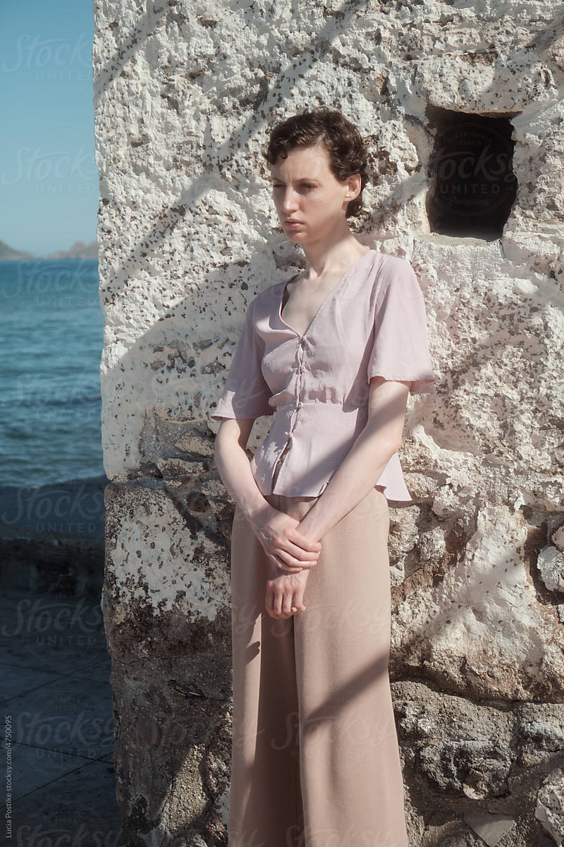 A woman in a pink blouse stands near a stone wall on the sea.