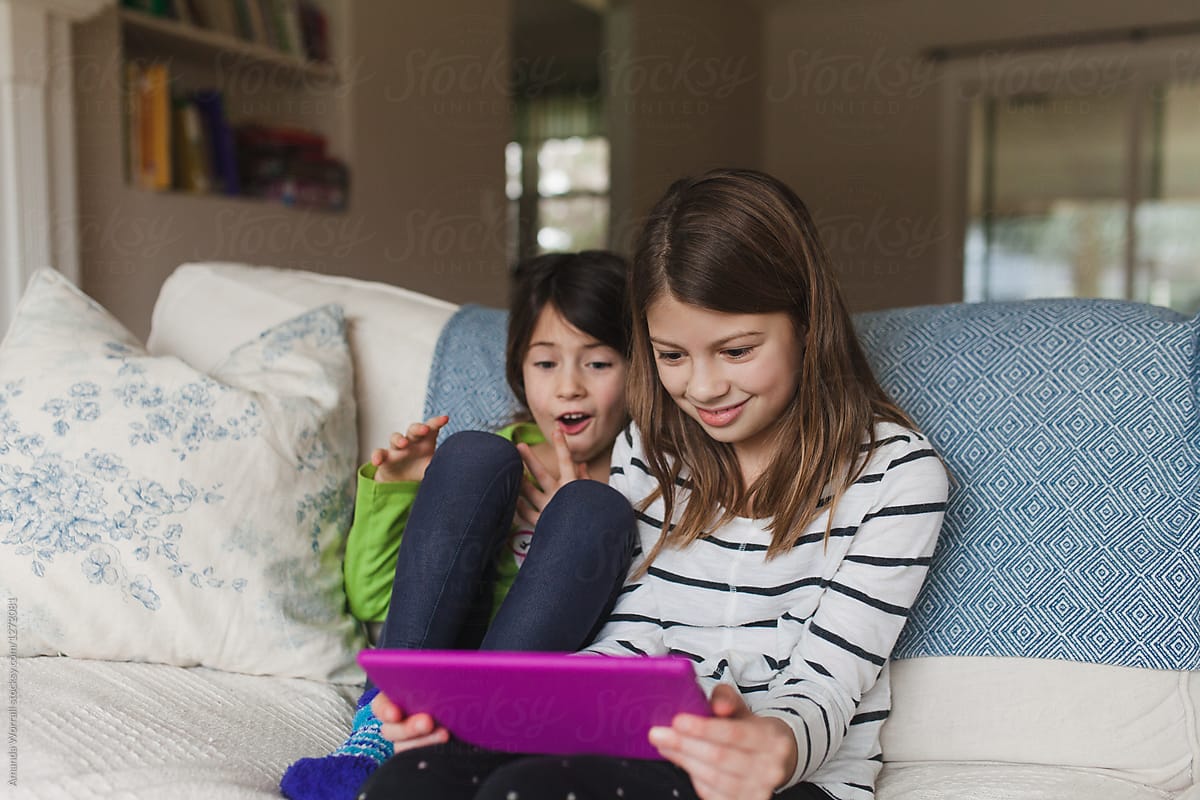 Two Girls Sitting On Sofa At Home Being Entertained By A Tablet By Stocksy Contributor