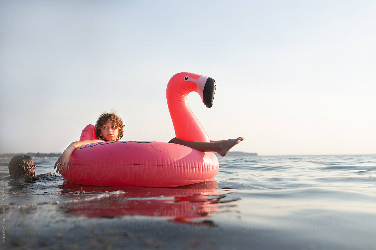 Boy floats in ocean on flamingo as faceless child swims up to him
