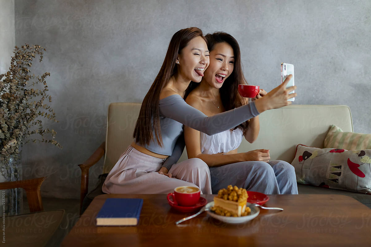 Two asian female Friends taking selfies together with coffee and cake sitting on a couch