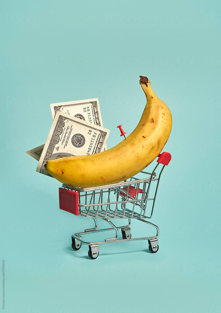 Shopping cart with dollar bills and banana on light blue background.