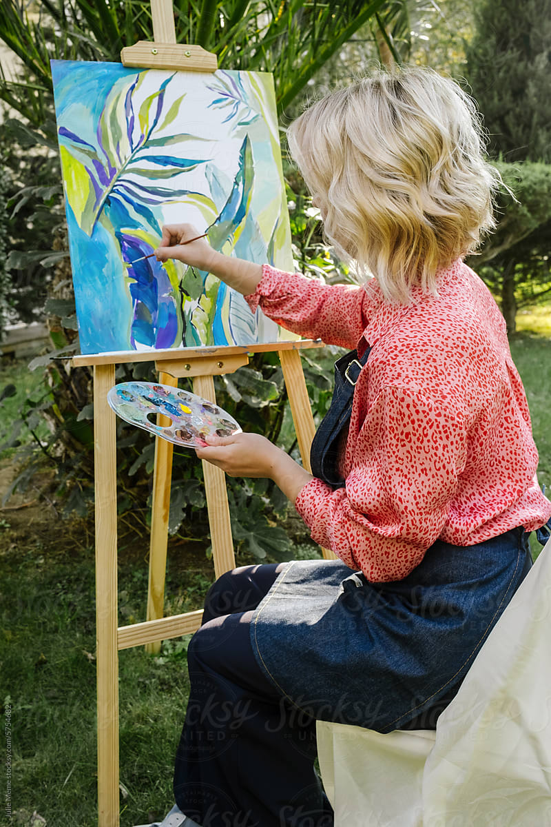 A blonde artist sits with her back turned, paints in nature