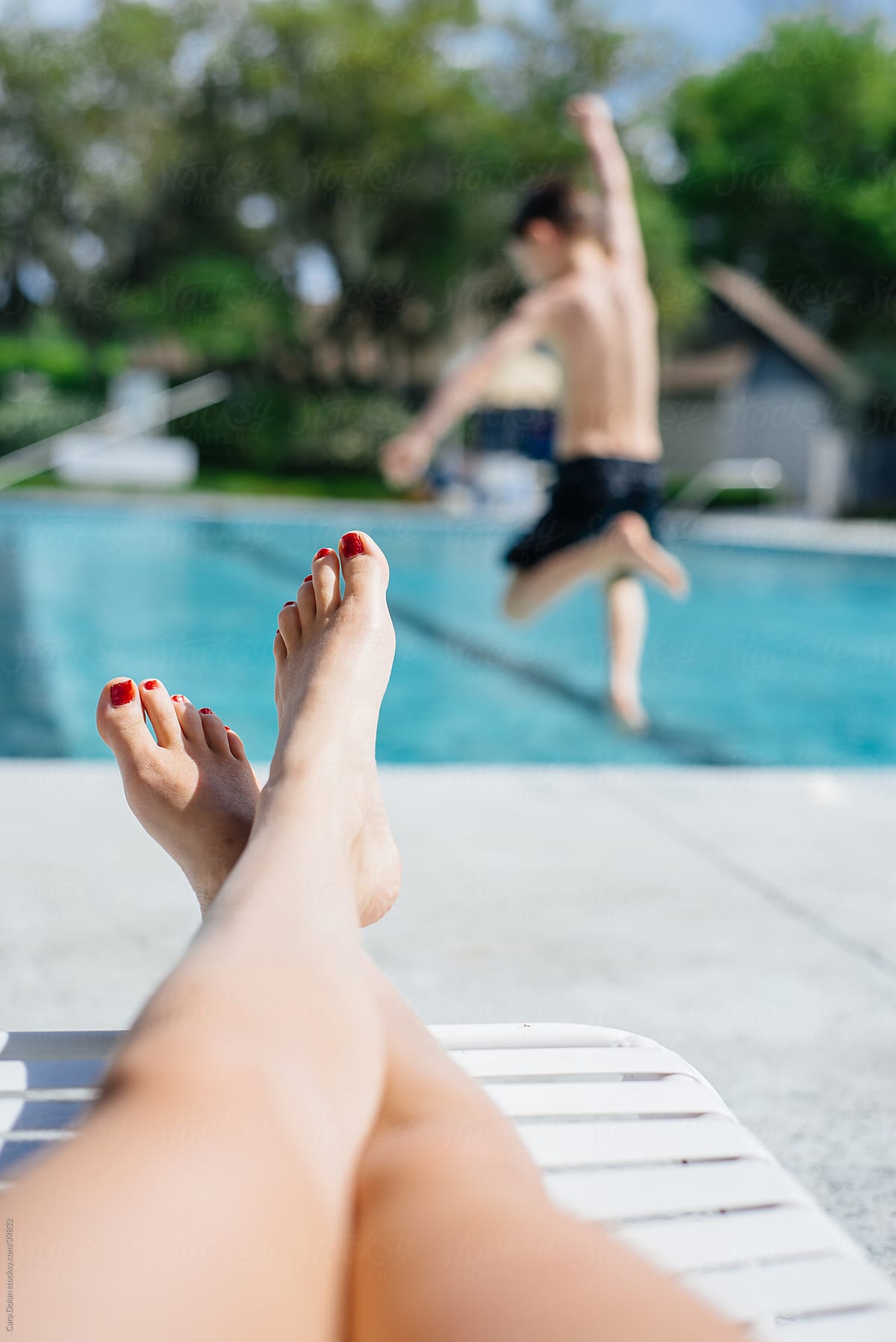 Mother relaxes poolside as her son leaps into the water