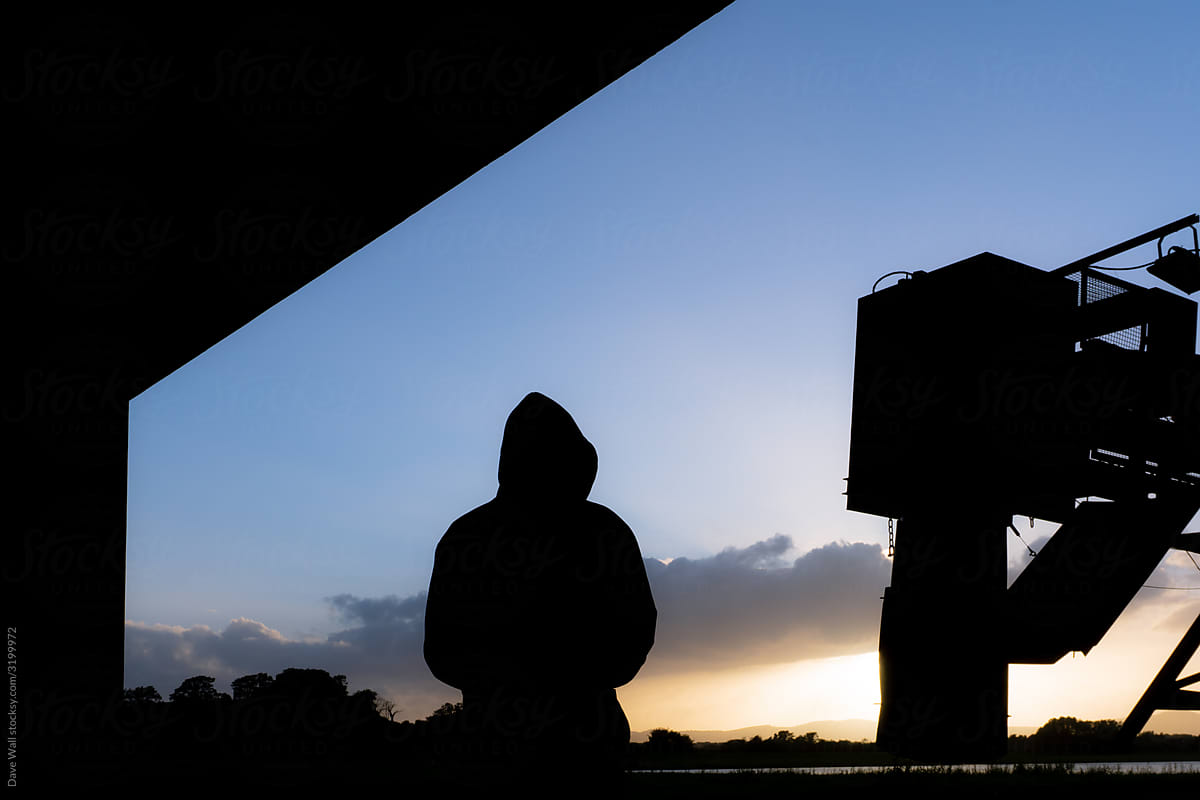 A hooded man silhouetted underneath a road bridge.