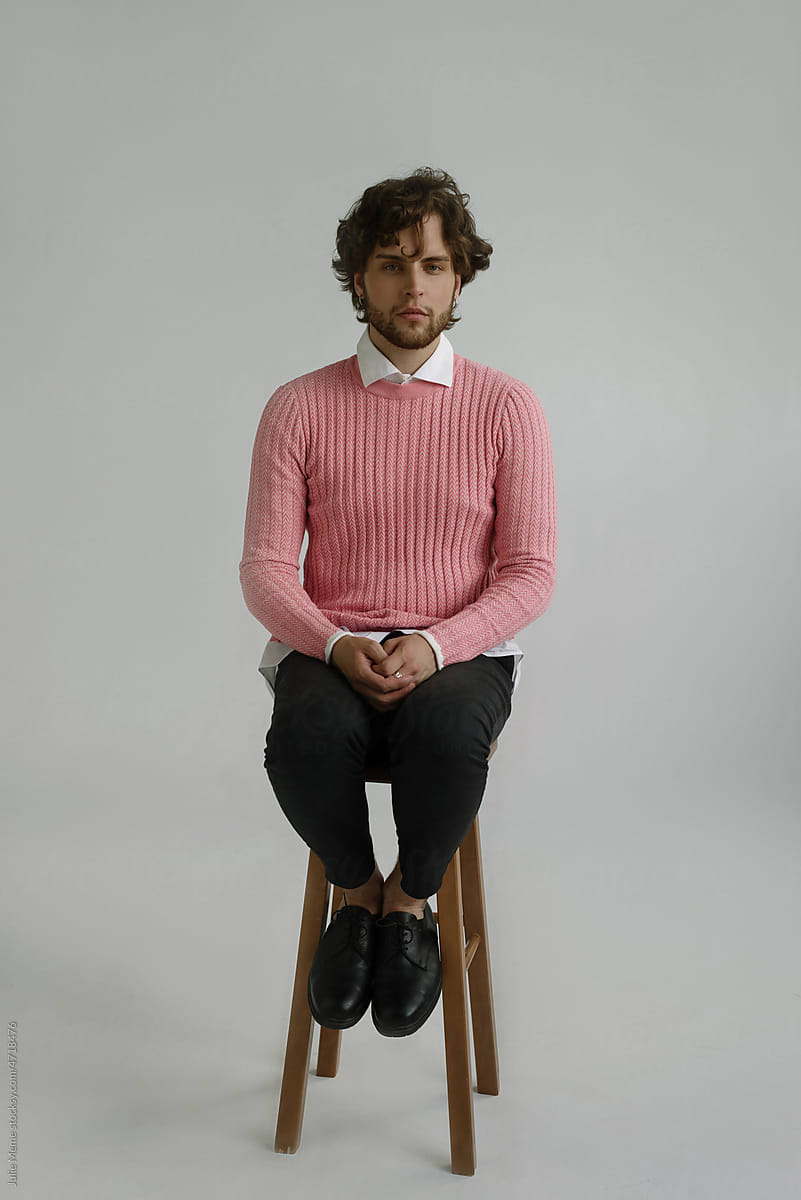 A stylish guy with curly hair sits in a pink pullover in the daylight