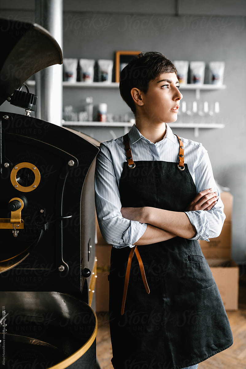 Brunette girl wearing apron leaning on big coffee machine.