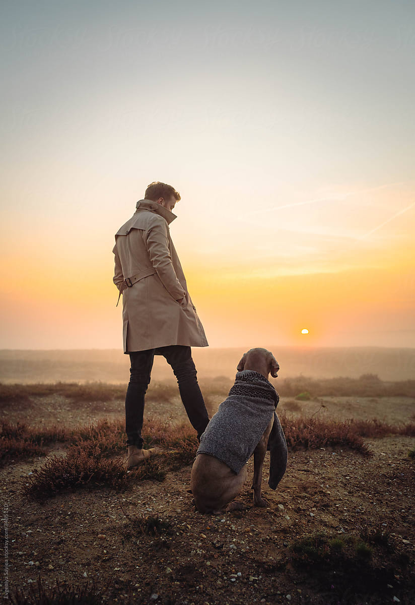 A young man in a beige coat standing in the foggy field with a dog by side at the sunrise