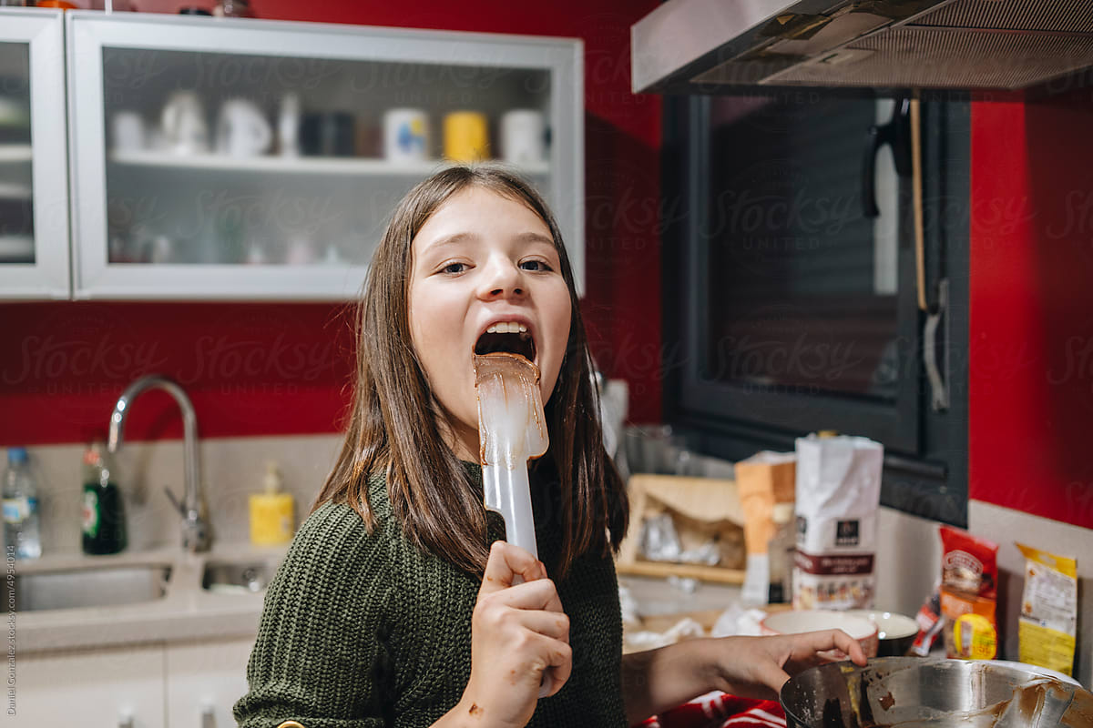 Funny girl licking dough from spatula in kitchen