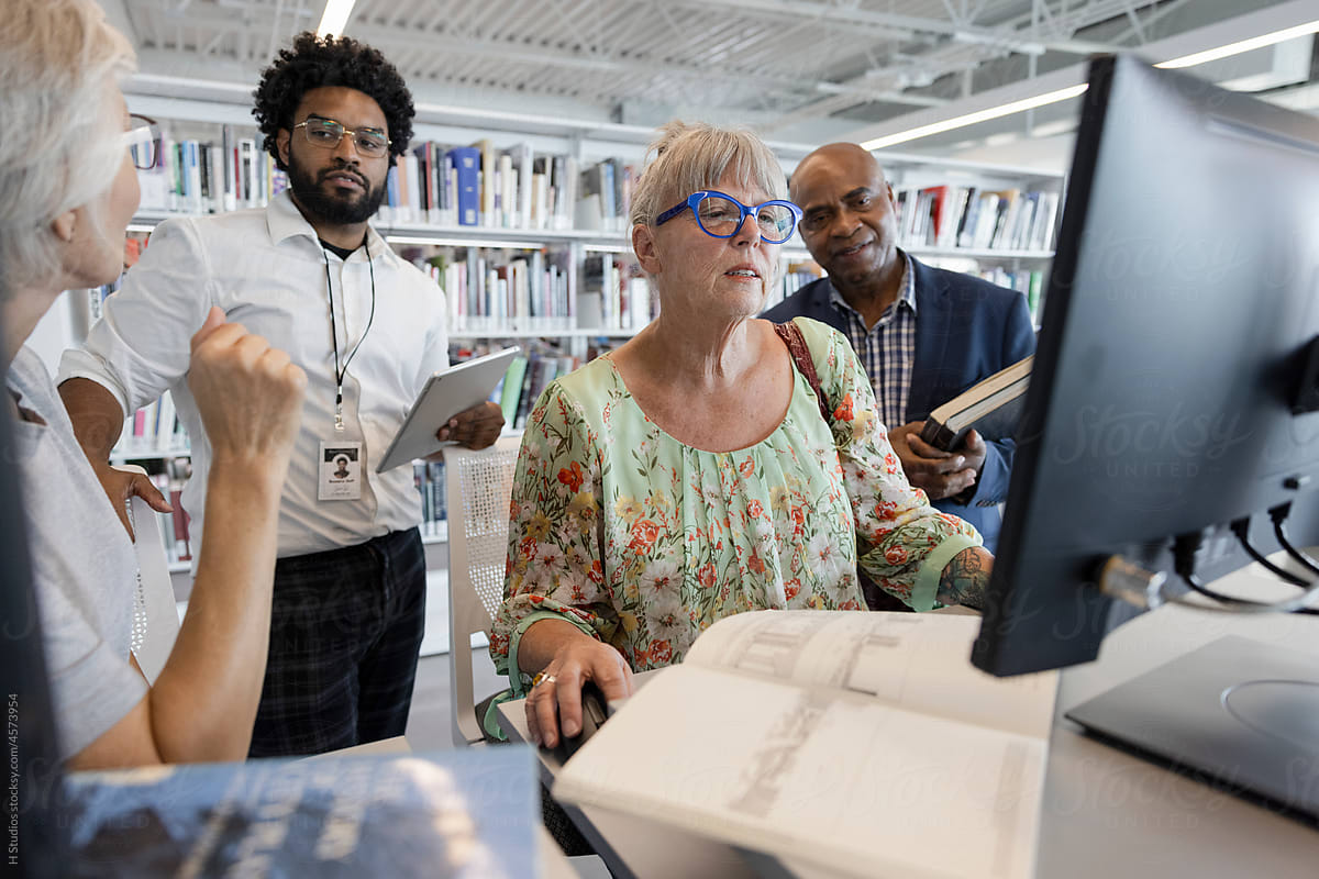 Male librarian helping senior woman at computer in library.