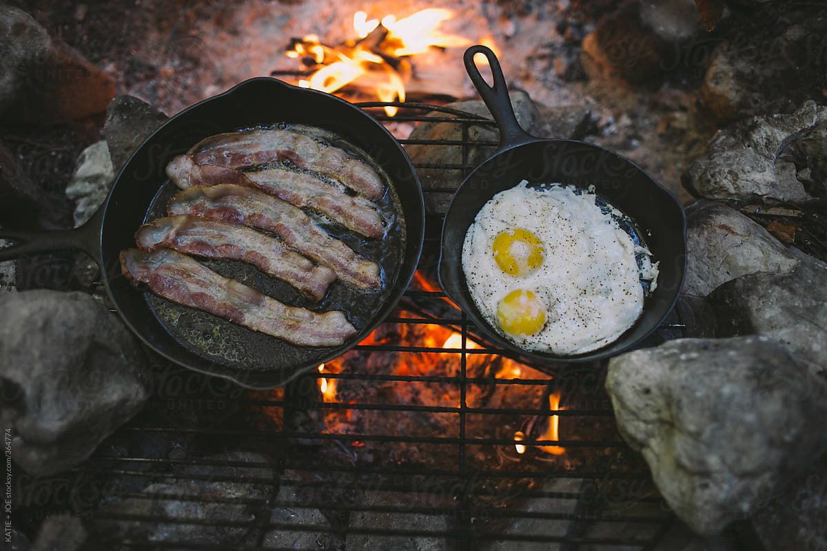 Bacon and eggs cooking on skillets over a fire