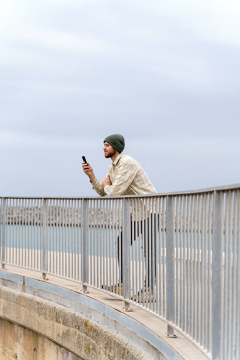 Modern man with smartphone leaning on bridge railing