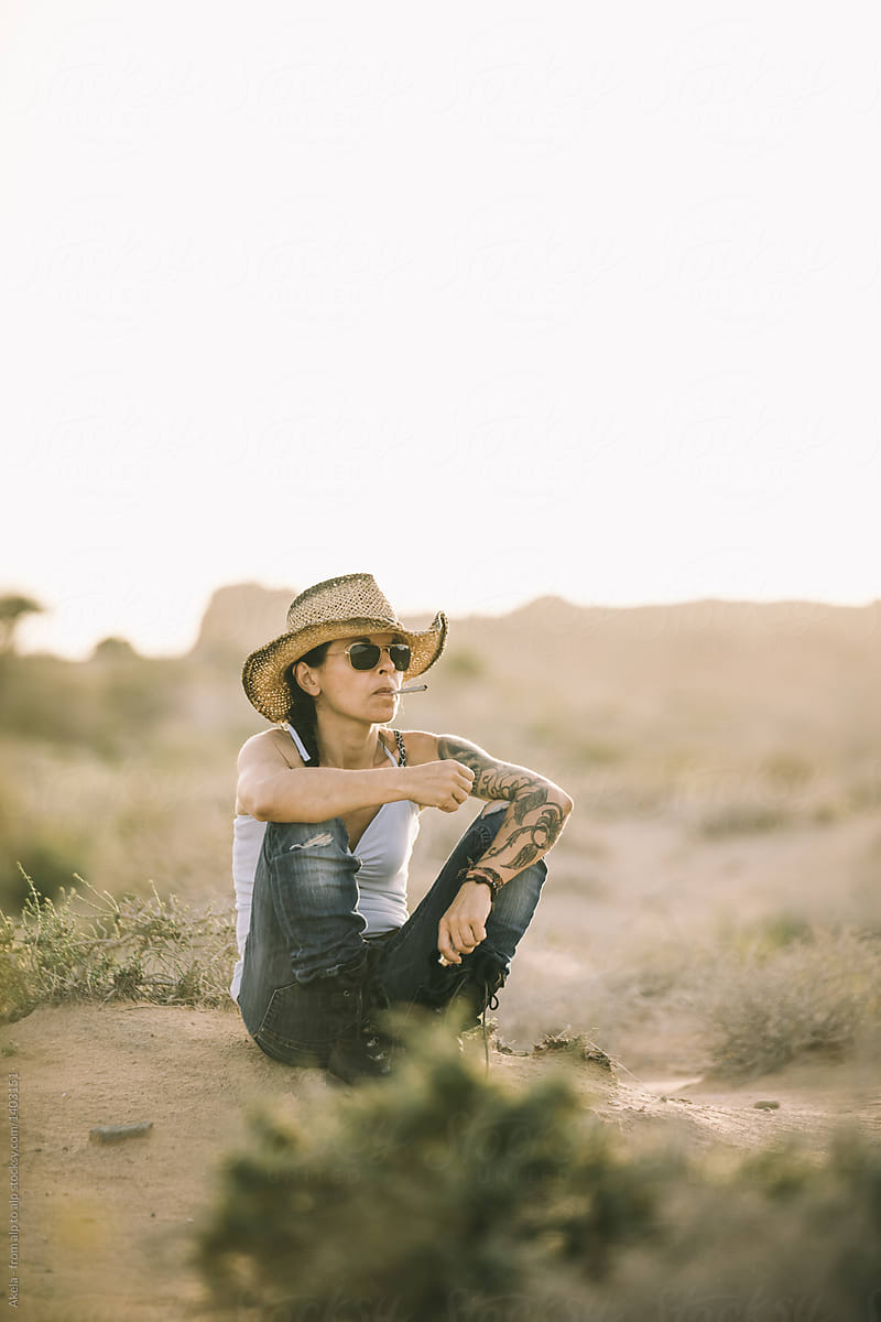 Casual Tattoed Woman With Cowboy Hat Sitting In Desert Smoking A Cigarette By Stocksy