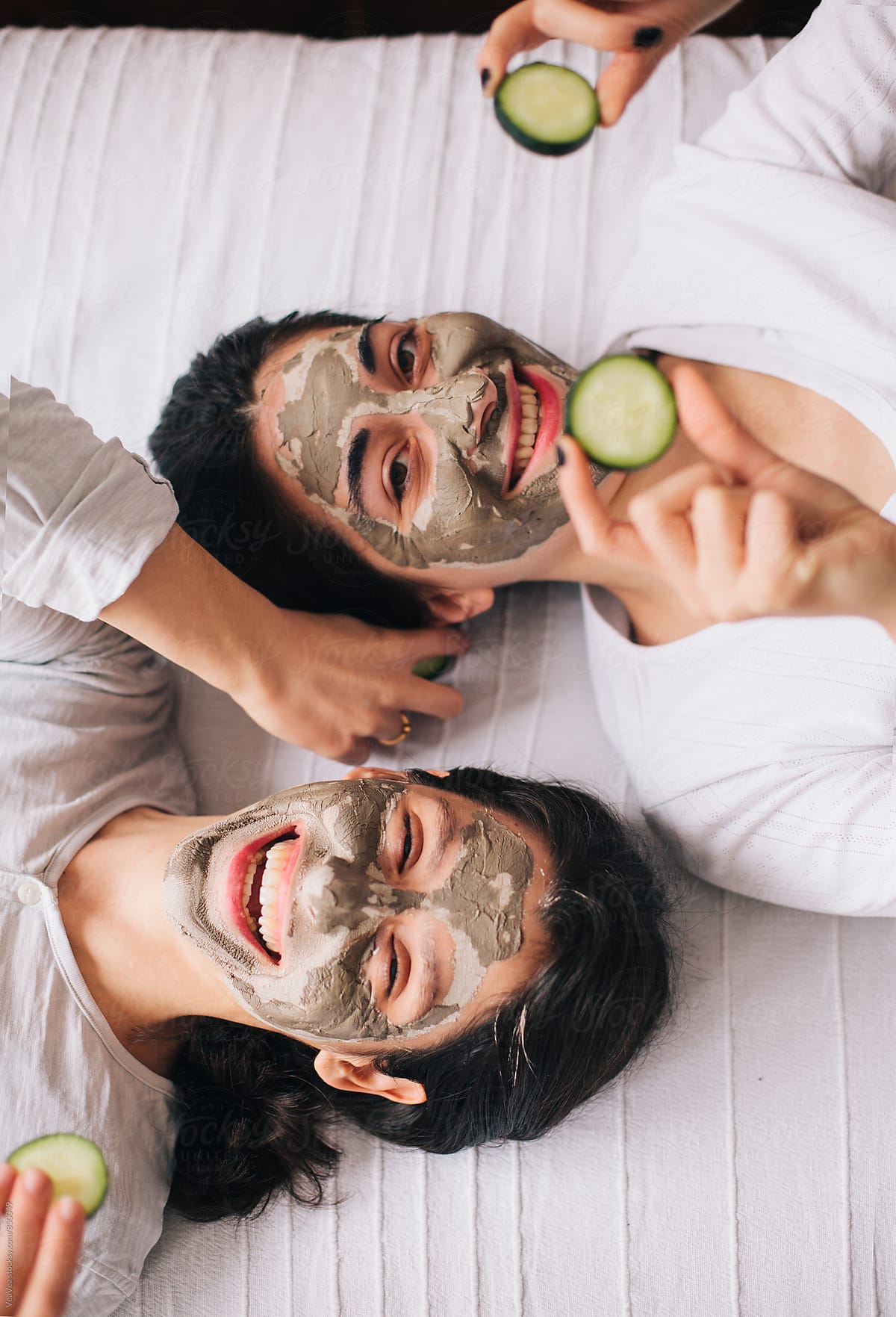 Two beautiful happy female friends with facial masks lying on the bed