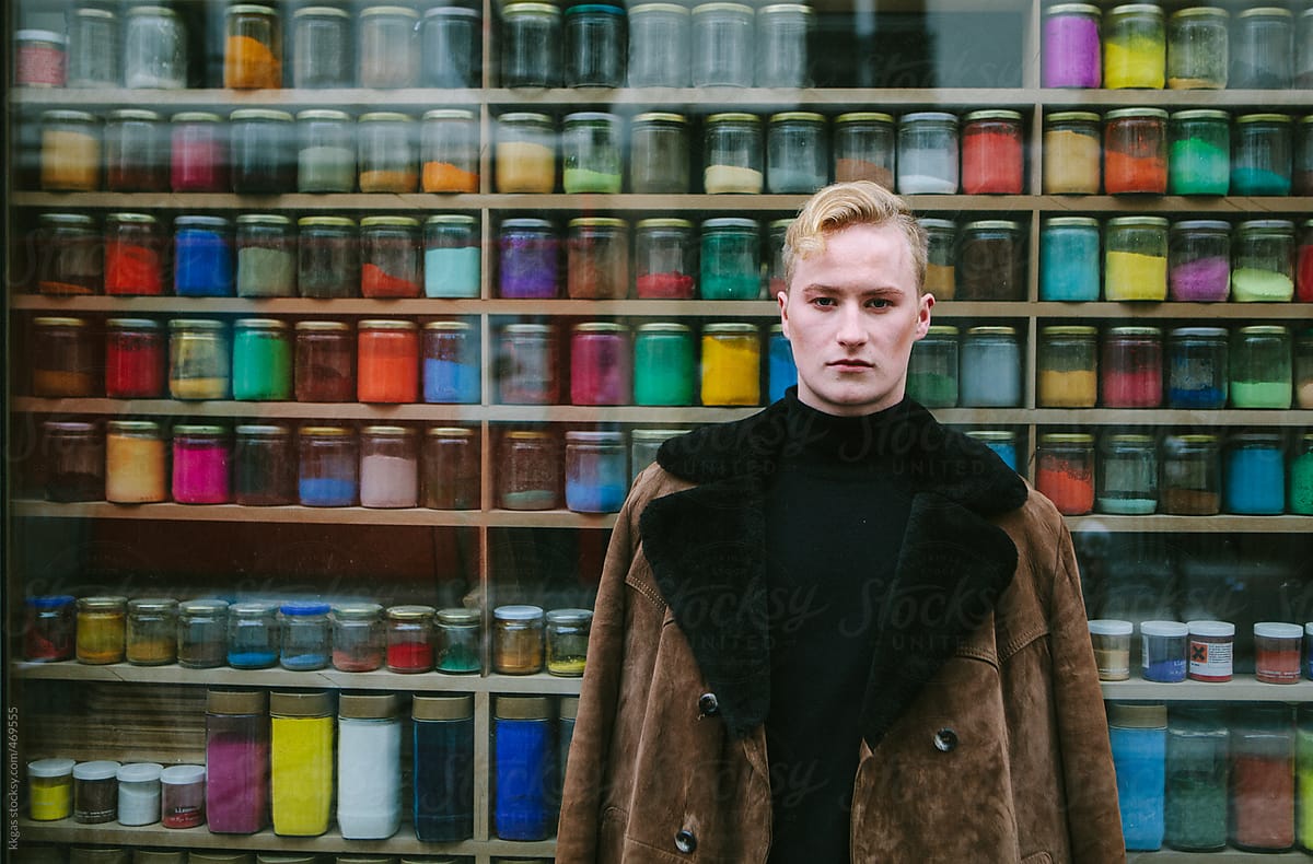 Blonde man next to a shop window filled with artists powder paints of many colours.