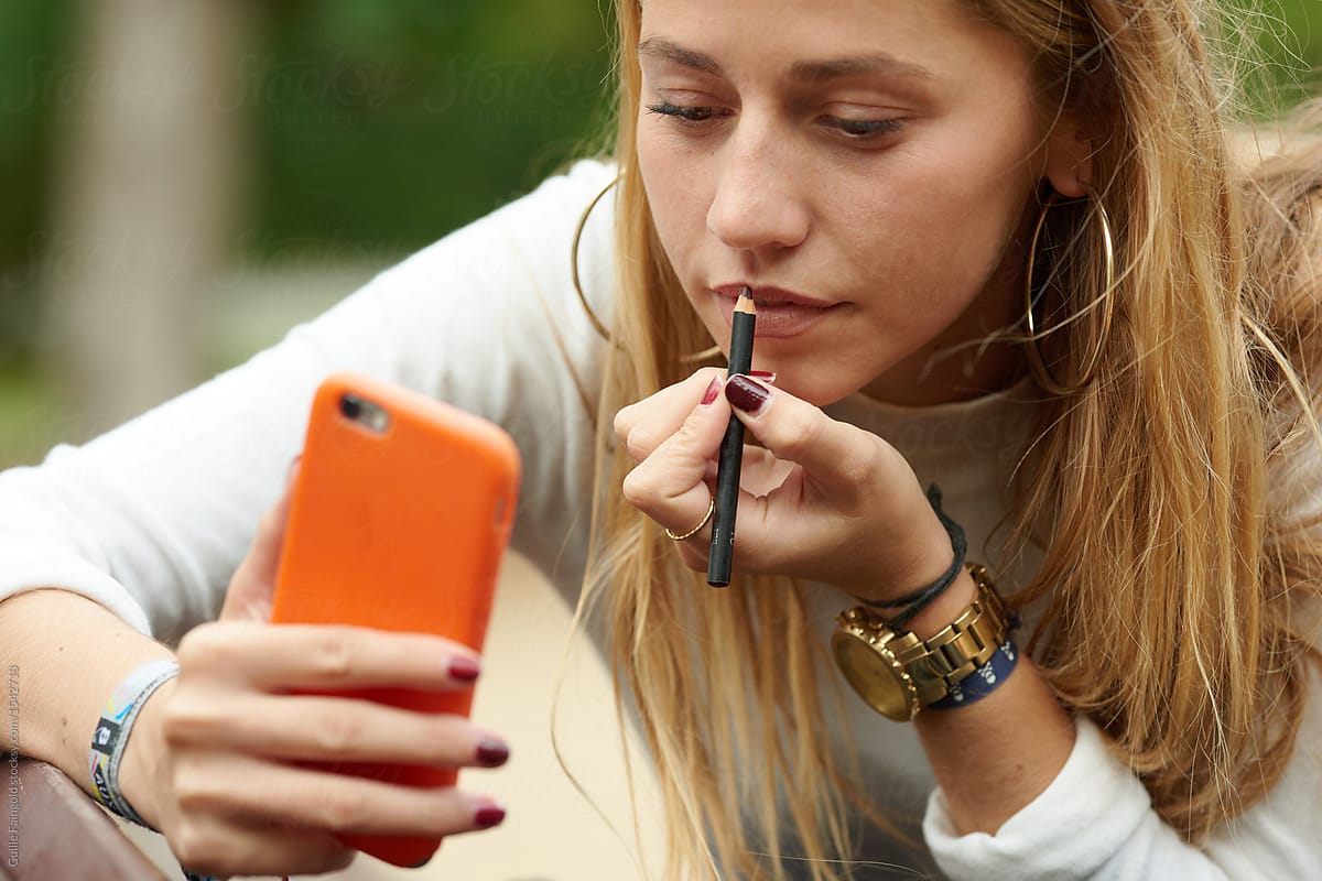 Blonde girl applying make-up with lip pencil and phone.