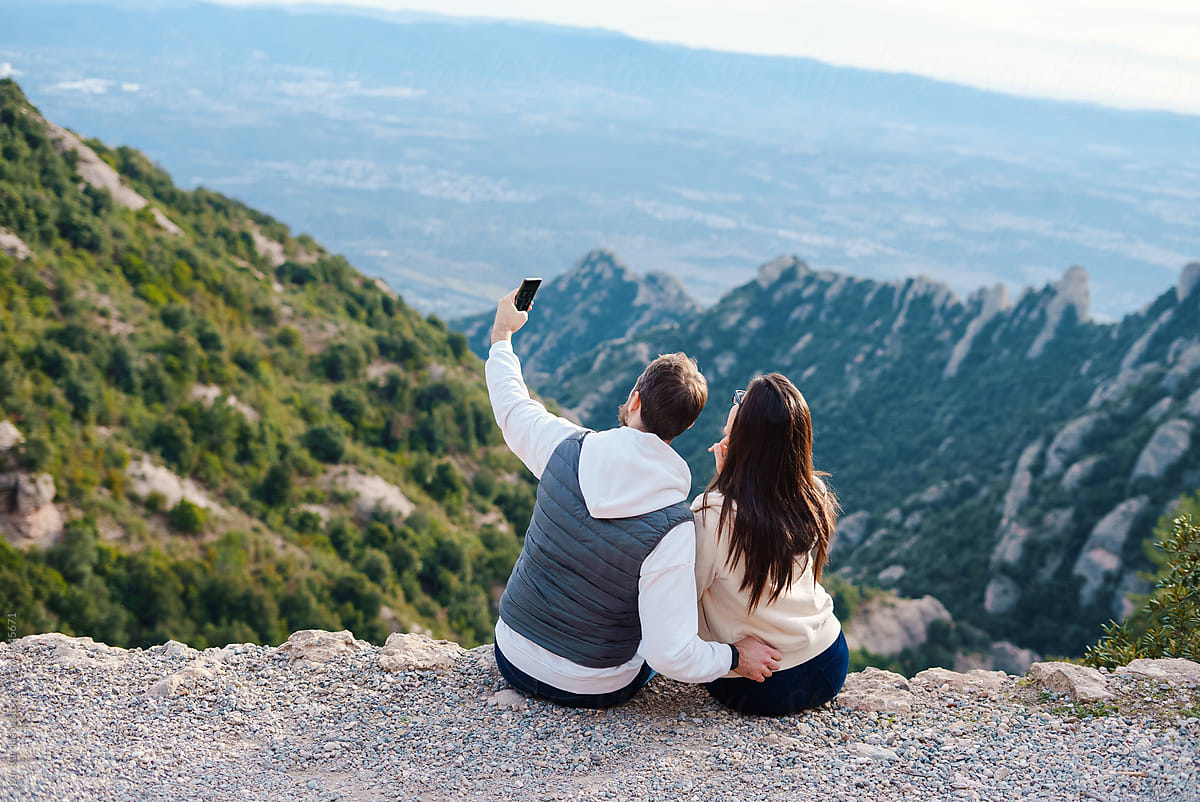 Couple sitting on rock peak in mountains taking selfie