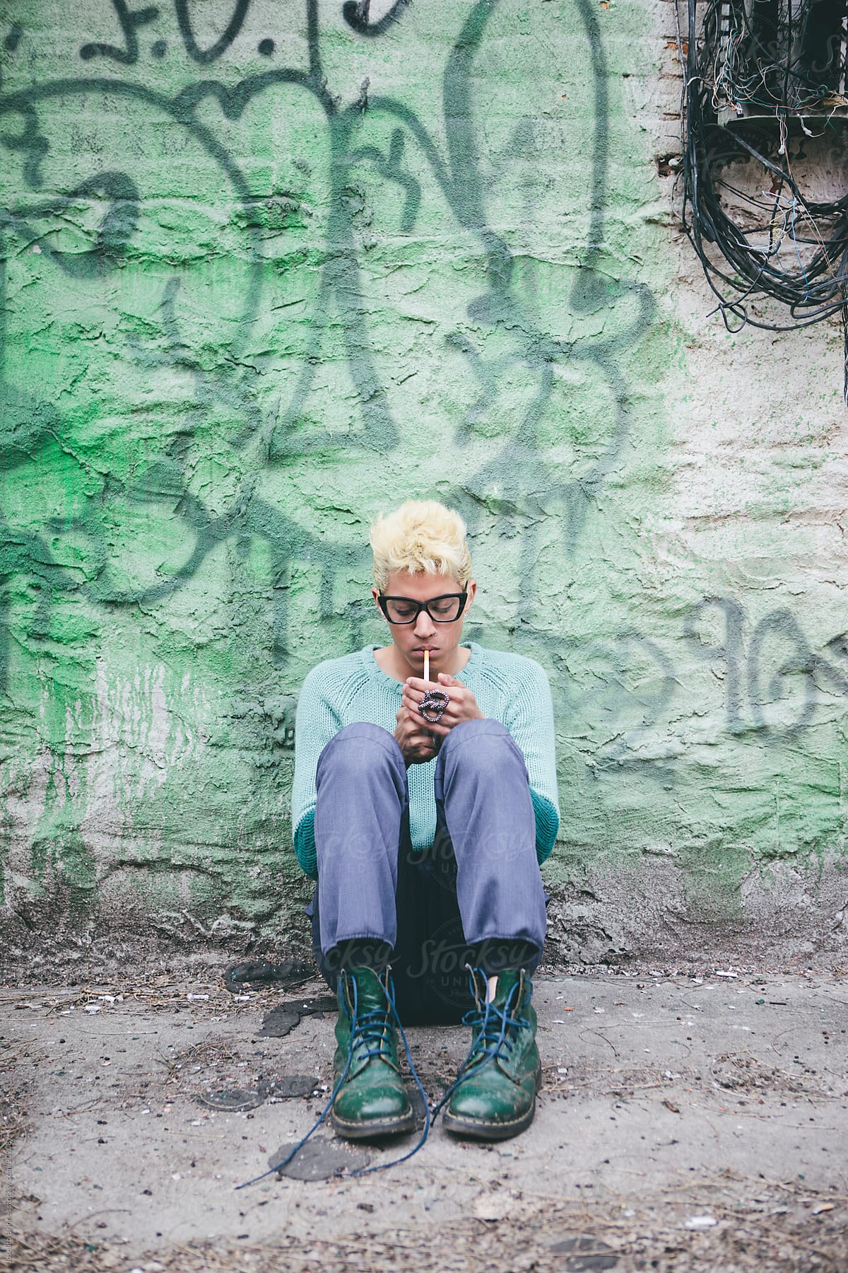 Stylish Young Man with Bleached Blond Hair in Spring and Summer Fashion Hiding in a Backyard while Smoking a Cigarette