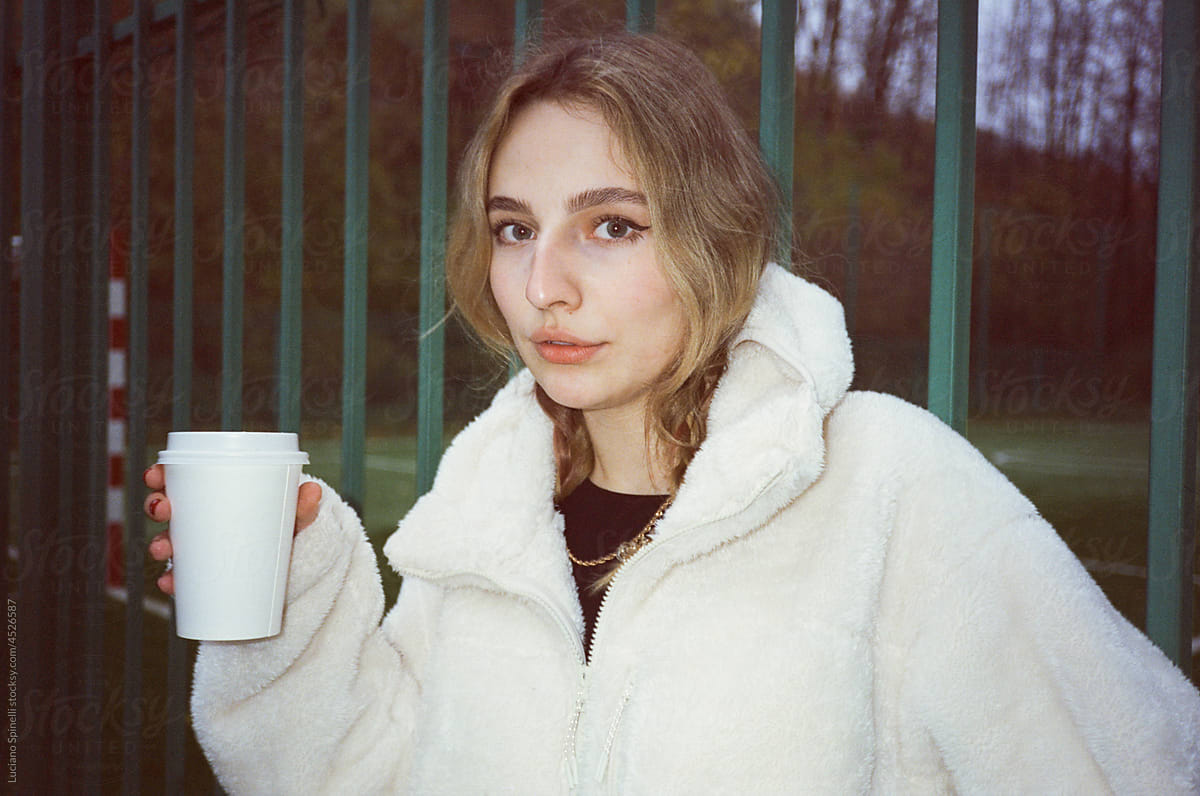 Woman in a white hoodie holding a cup of tea next to a green fence