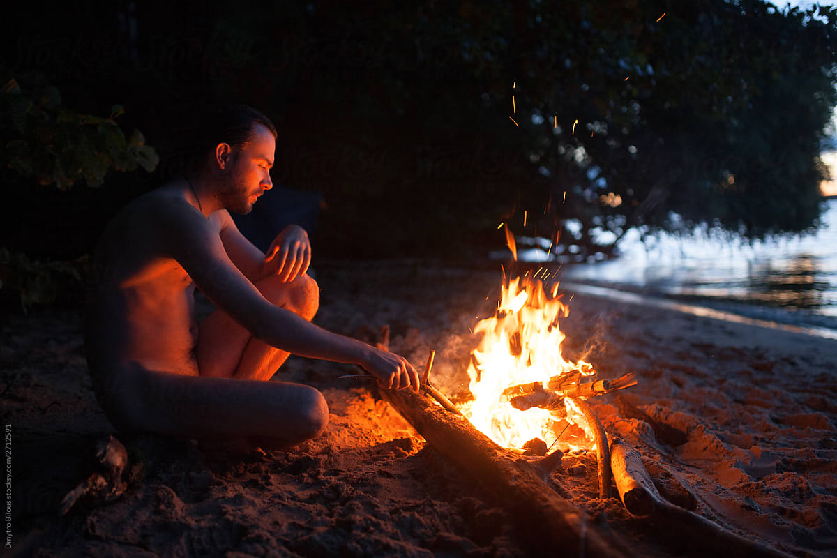 naked guy sitting by the fire on a log on the beach