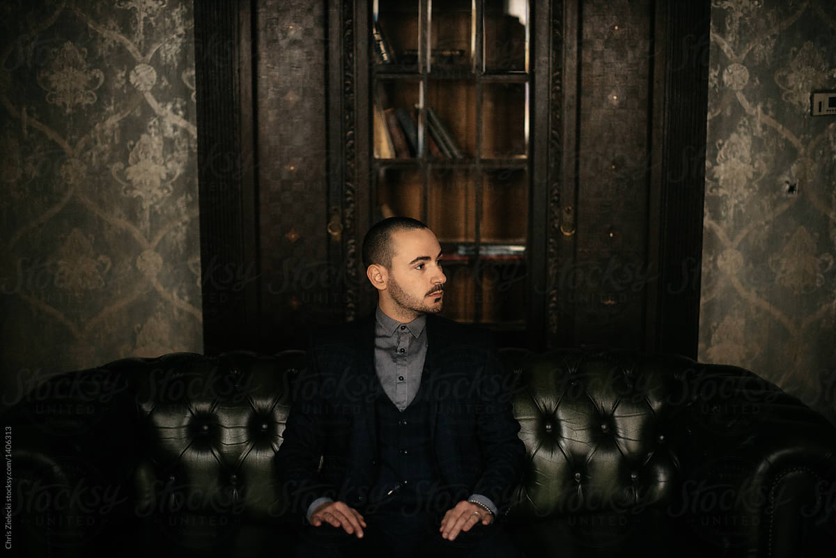 Stylish groom in suit sitting on sofa in dark room