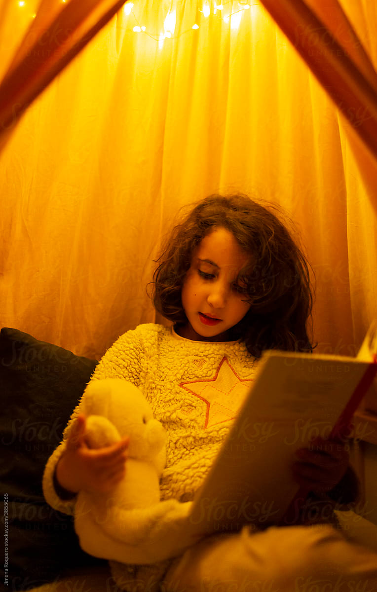 Kid reading book in bed with teddy bear under fairy lights
