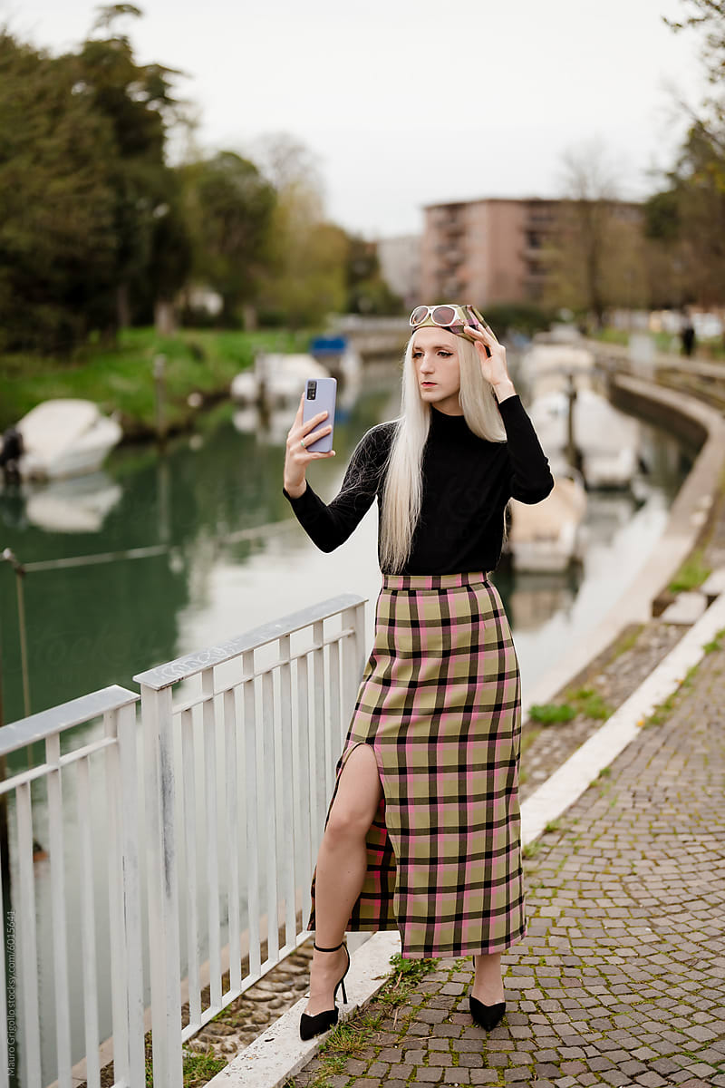 A stylish blond man using a smartphone for a selfie along a river